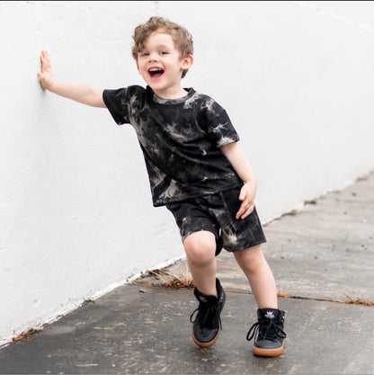 Toddler with Grunge Tie Dye Shorts and Tshirt in urban setting leaning against a white wall with sneakers on